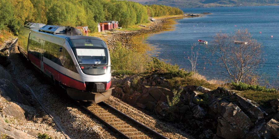 A silver Nordland train travels past serene blue water and small red fisherman’s huts.  