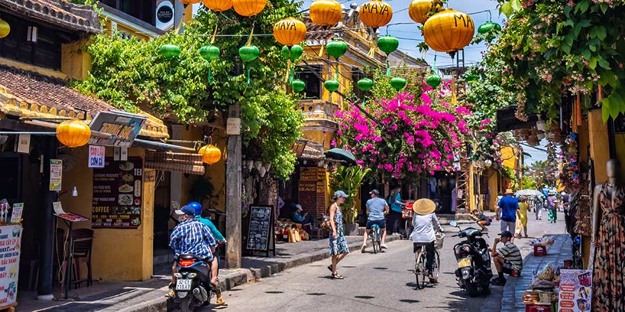 A vibrant street in Hoi An full of colourful buildings and flowers, decorative lanterns hung up above, and pedestrians and motorcyclists buzzing by on the busy street. 