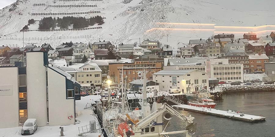 Honningsvag harbour covered in snow, a white hill can be seen behind some of the town’s houses. 